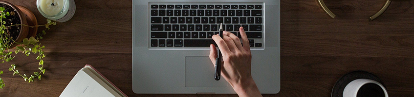 Schreibtisch von oben herab fotografiert, Ausschnitt eines Laptops, einer Hand die mit einem Stift auf den Monitor des Laptops zeigt